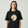 A young woman wearing a classic black short-sleeved t-shirt. The shirt features a large white daisy graphic in the center with a yellow middle, missing one petal at the bottom left. Above the flower, the word "PEACEMINUSONE" is printed in white text with a small lightning bolt logo